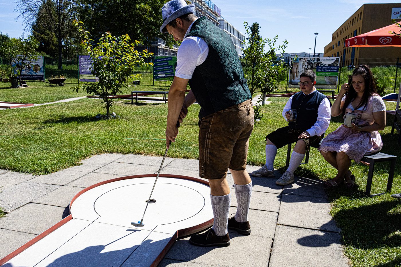 Minigolfspieler beim Einlochen, zwei Gäste schauen von der Bank aus zu